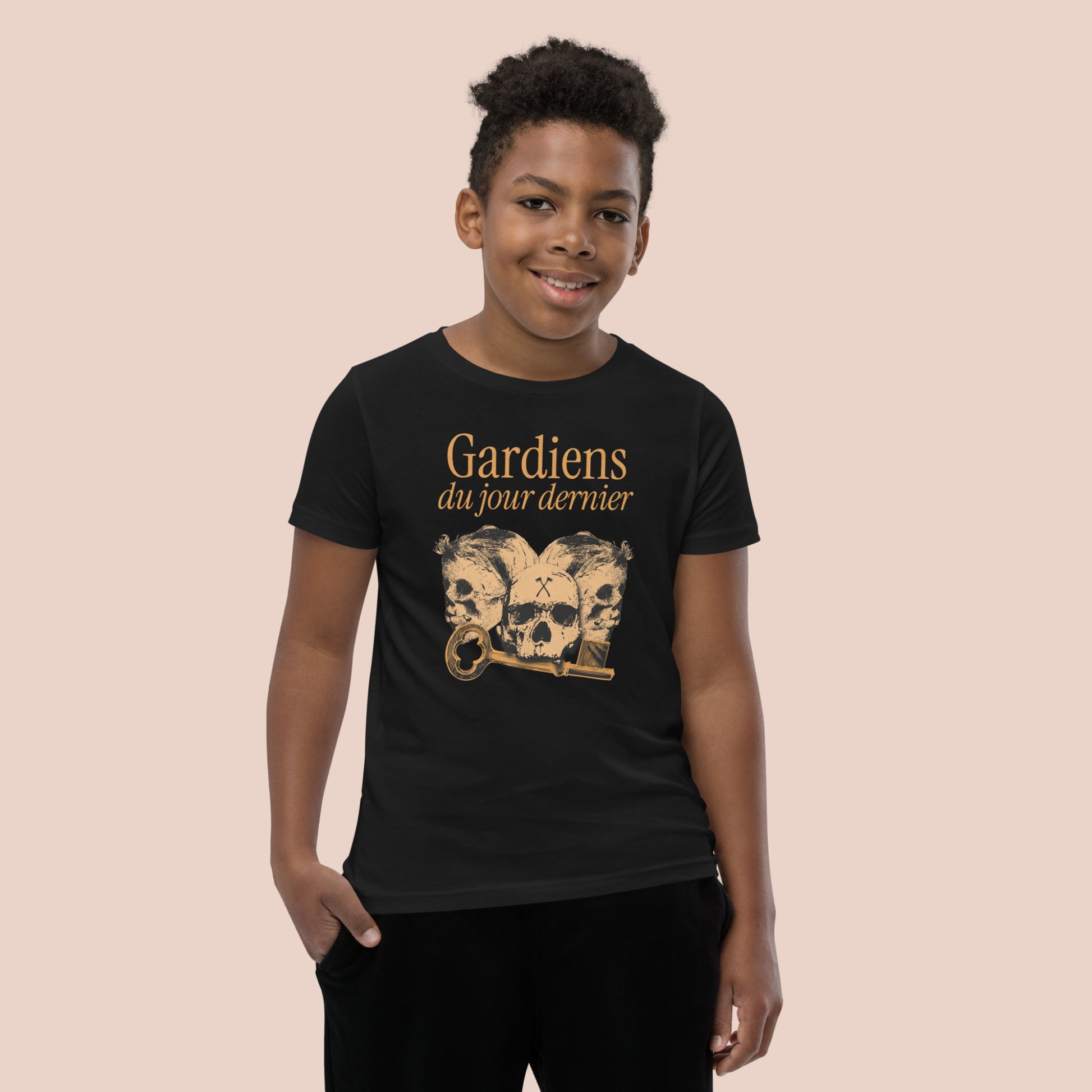 Garçon souriant portant un t-shirt noirl illustré de têtes de squelette et inscrit Gardiens du jour dernier. Vue du devant.